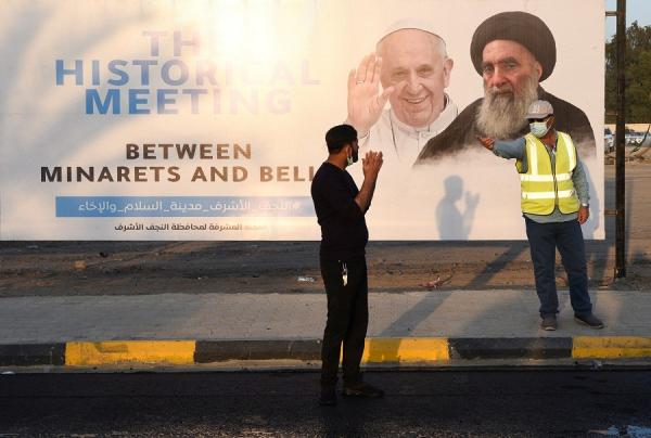 Dua pria berdiri di dekat poster pertemuan Paus Fransiskus dan Ayatollah Ali al-Sistani di salah satu kota Irak. (Foto: Reuters)